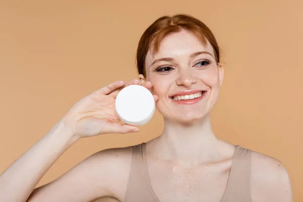 Pleased redhead woman holding cosmetic cream isolated on beige — Stock Photo