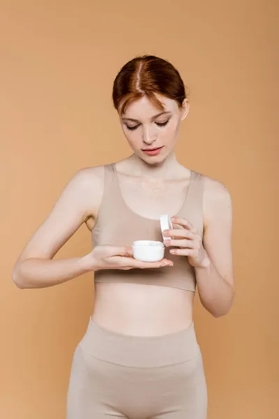 Femme taches de rousseur en haut regardant la crème cosmétique isolée sur beige — Photo de stock