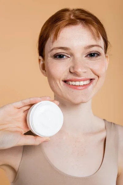 Retrato de mujer pecosa sonriente sosteniendo crema y mirando a la cámara aislada en beige - foto de stock