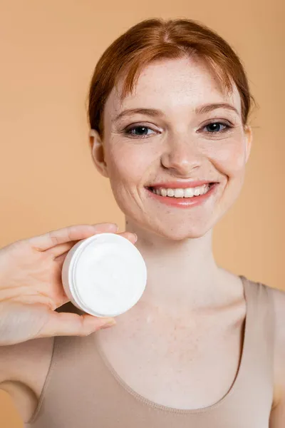 Retrato de mulher sardenta sorrindo segurando creme e olhando para a câmera isolada no bege — Fotografia de Stock