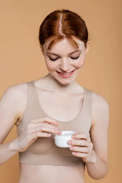 Young redhead woman smiling while holding cosmetic cream isolated on beige — Stock Photo