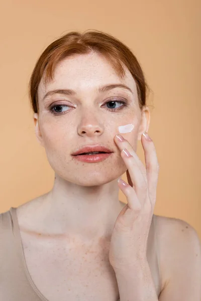 Ritratto di giovane donna che applica crema cosmetica sul viso isolato sul beige — Foto stock