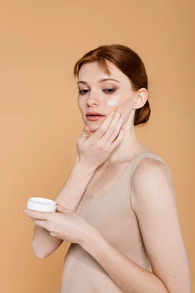 Mujer pecosa y pelirroja aplicando crema en la mejilla aislada en beige - foto de stock