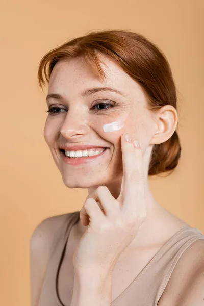Femme aux cheveux rouges positifs appliquant de la crème sur la joue isolée sur beige — Photo de stock