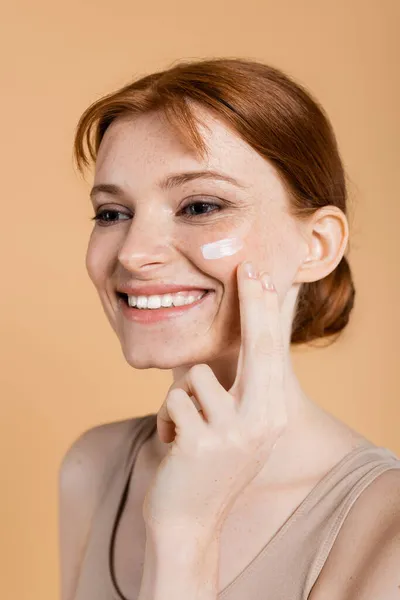 Mulher ruiva positiva aplicando creme na bochecha isolada no bege — Fotografia de Stock