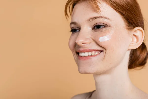 Lächelnde rothaarige Frau mit perfekter Haut und Creme im Gesicht, die isoliert auf Beige wegschaut — Stockfoto