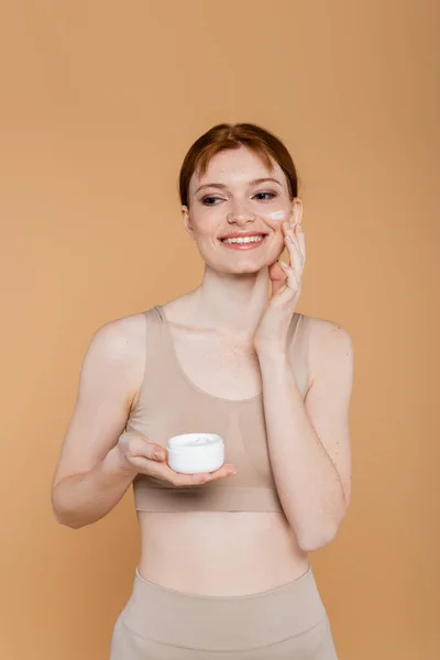 Jolie femme aux cheveux rouges appliquant de la crème cosmétique sur le visage isolé sur beige — Photo de stock