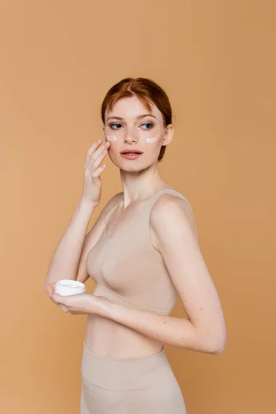 Jeune femme en haut appliquant de la crème cosmétique sur les joues isolées sur beige — Photo de stock