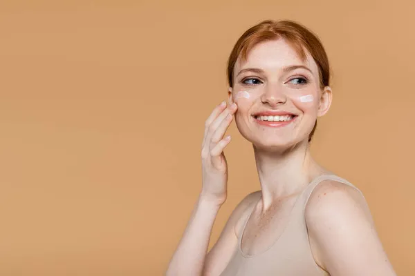 Mujer de pelo rojo positivo aplicando crema cosmética en las mejillas aisladas en beige - foto de stock