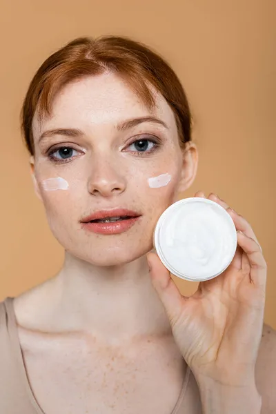 Young redhead woman showing container with cosmetic cream isolated on beige — Stock Photo