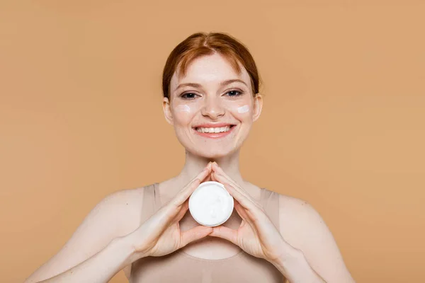 Mujer pelirroja sonriente sosteniendo crema cosmética y mirando a la cámara aislada en beige - foto de stock