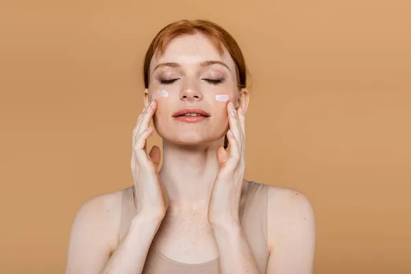 Freckled redhead woman with closed eyes applying cream on cheeks isolated on beige — Stock Photo