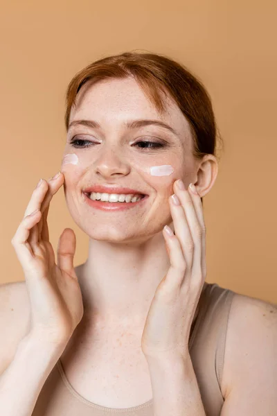 Mulher de cabelos vermelhos alegres aplicando creme cosmético em bochechas isoladas em bege — Fotografia de Stock