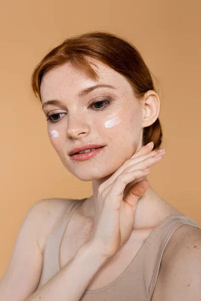 Jolie femme aux cheveux rouges avec crème cosmétique sur le visage posant isolée sur beige — Photo de stock