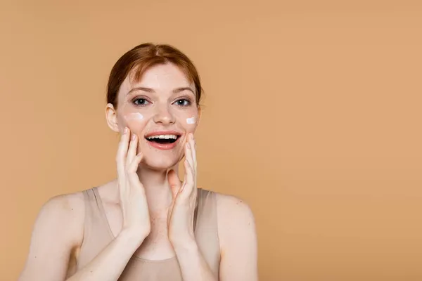 Excited red haired woman with cosmetic cream touching cheeks isolated on beige — Stock Photo