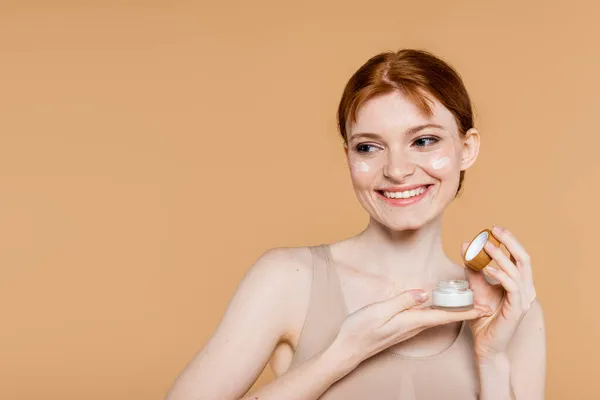 Mujer feliz sosteniendo contenedor con crema cosmética y mirando hacia otro lado aislado en beige - foto de stock