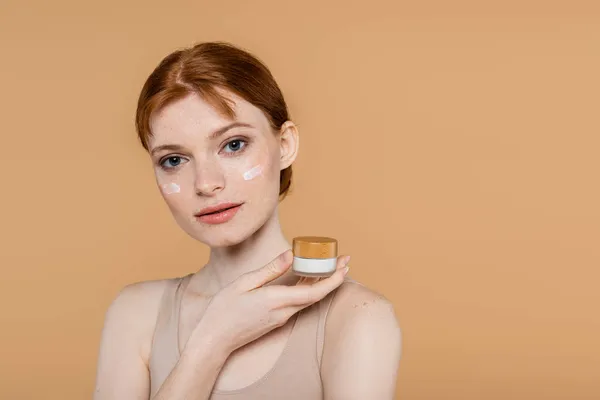 Pretty young woman with cream on face holding jar isolated on beige — Stock Photo