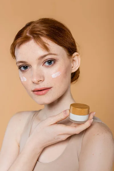 Jeune femme avec des taches de rousseur tenant pot avec de la crème tout en posant isolé sur beige — Photo de stock