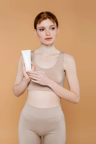 Jolie femme aux cheveux rouges tenant tube avec crème cosmétique isolée sur beige — Photo de stock