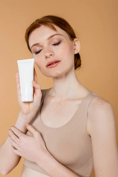 Mujer joven pelirroja con los ojos cerrados sosteniendo tubo con loción cosmética aislada en beige - foto de stock