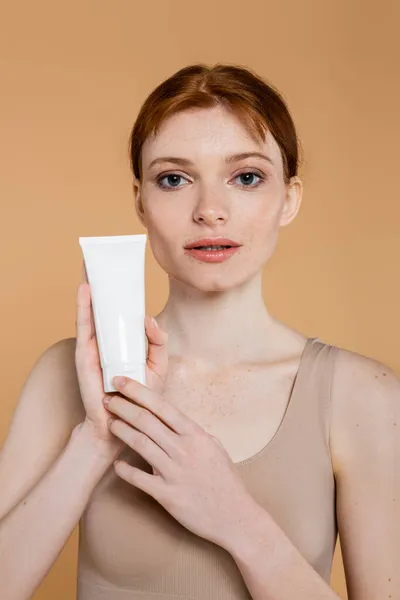 Jolie femme aux cheveux rouges tenant tube avec crème cosmétique et regardant la caméra isolée sur beige — Photo de stock