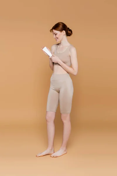Vista lateral de mujer descalza sonriente sosteniendo tubo con loción cosmética sobre fondo beige - foto de stock