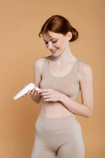Femme aux cheveux rouges souriante regardant tube avec crème cosmétique isolé sur beige — Photo de stock