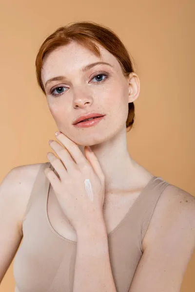 Red haired woman with cream on hand looking at camera isolated on beige — Stock Photo