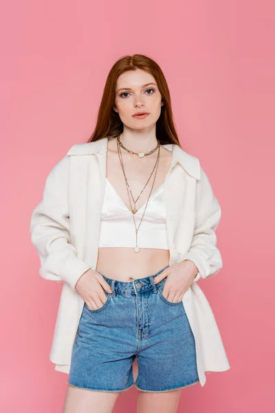 Young red haired woman in jacket holding hands in pockets of shorts isolated on pink — Stock Photo