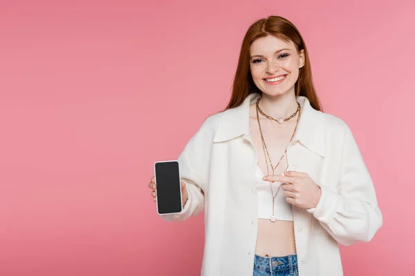 Jolie rousse femme en veste pointant vers smartphone isolé sur rose — Photo de stock