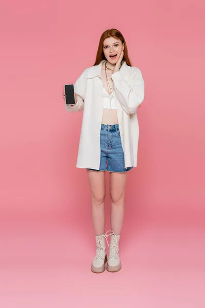 Excited woman in jacket and necklaces holding cellphone with blank screen on pink background — Stock Photo