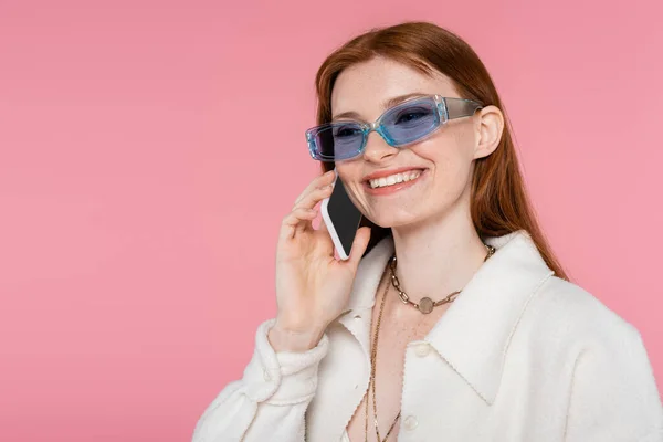 Elegante donna dai capelli rossi in occhiali da sole che parla su smartphone isolato su rosa — Foto stock