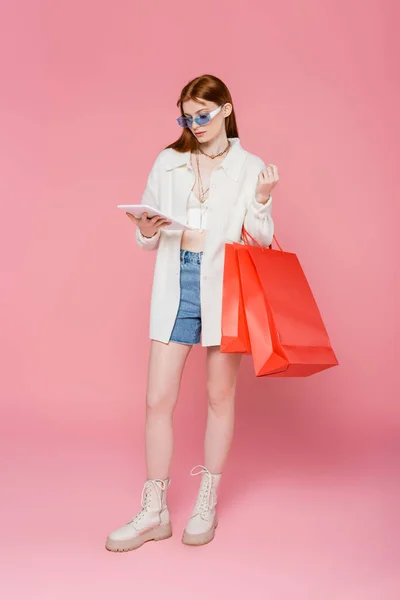 Stylish red haired woman holding digital tablet and shopping bags on pink background — Stock Photo