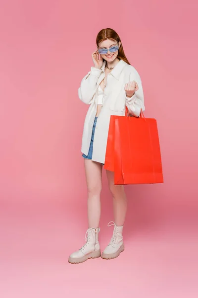 Mulher sorridente em óculos de sol olhando para sacos de compras vermelhos em fundo rosa — Fotografia de Stock