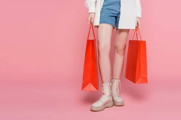 Vista recortada de la mujer en pantalones cortos de mezclilla sosteniendo bolsas de compras sobre fondo rosa - foto de stock