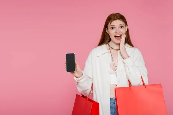 Mulher de cabelos vermelhos animado com sacos de compras segurando smartphone com tela do banco isolado em rosa — Fotografia de Stock