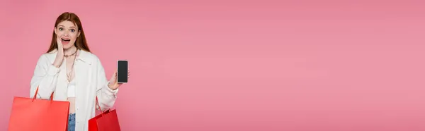 Femme rousse excitée avec des sacs à provisions tenant le téléphone portable avec écran vide isolé sur rose, bannière — Photo de stock
