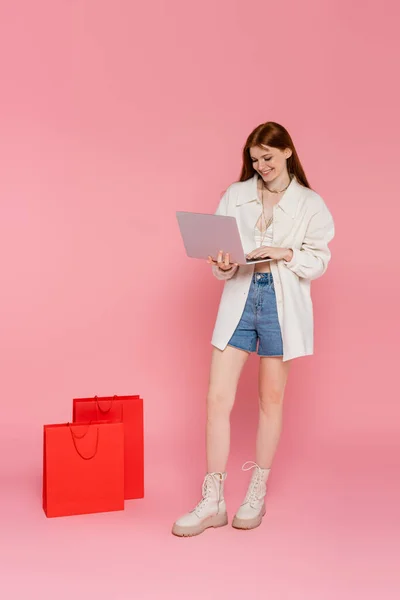 Позитивна червона жінка використовує ноутбук біля сумки на рожевому фоні — стокове фото