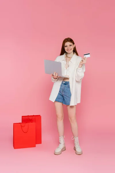 Sonriente mujer pelirroja sosteniendo tarjeta de crédito y portátil cerca de bolsas de compras en fondo rosa - foto de stock