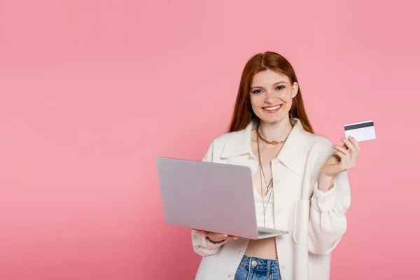 Elegante donna dai capelli rossi in possesso di carta di credito e laptop isolato su rosa — Foto stock