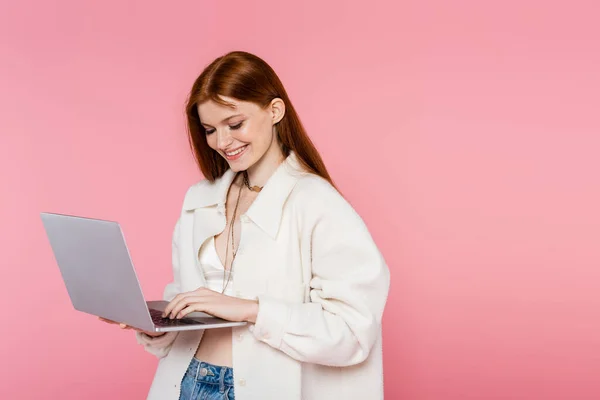 Smiling red haired woman in jacket using laptop isolated on pink — Stock Photo