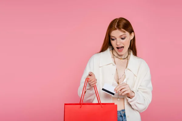 Дивовижна руда жінка тримає кредитну картку і дивиться на торгову сумку ізольовано на рожевому — стокове фото