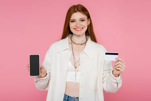 Mujer pelirroja alegre en chaqueta y collares con tarjeta de crédito y teléfono celular aislado en rosa - foto de stock