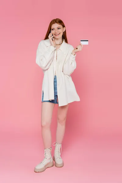 Longitud completa de mujer de pelo rojo elegante hablando en el teléfono móvil y la celebración de la tarjeta de crédito en el fondo rosa - foto de stock