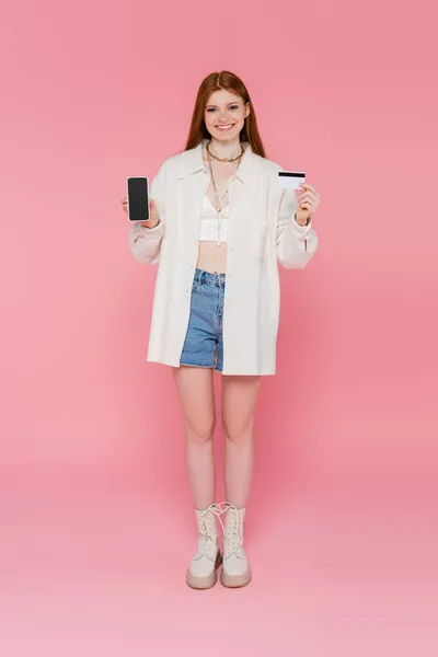 Красива і стильна руда волохата жінка тримає мобільний телефон і кредитну картку на рожевому фоні — стокове фото