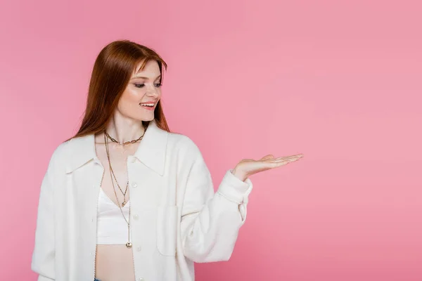 Mujer sonriente de pelo rojo en chaqueta apuntando con la mano aislada en rosa - foto de stock