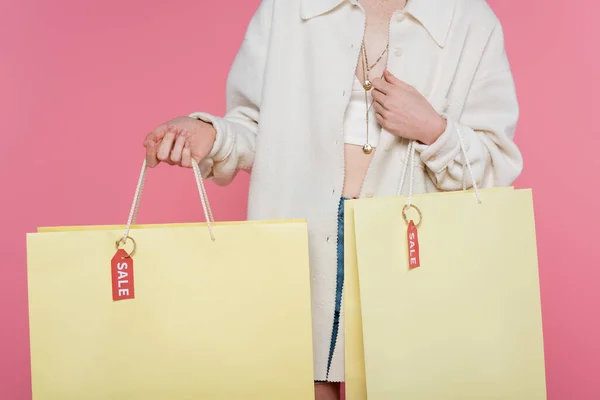 Vue recadrée d'une jeune femme élégante tenant des sacs à provisions avec étiquettes de prix et lettrage de vente isolé sur rose — Photo de stock