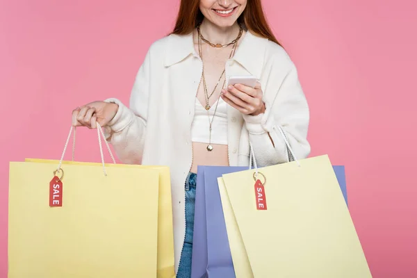 Vista recortada de la mujer con estilo con bolsas de compras utilizando teléfono inteligente aislado en rosa - foto de stock