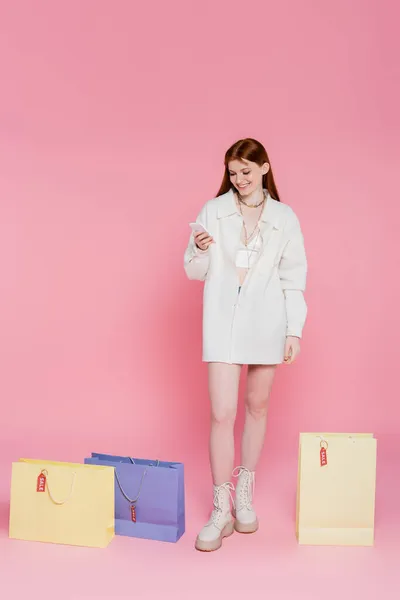 Shopaholic sorrindo usando smartphone perto de sacos de compras com etiquetas de preço e venda lettering em fundo rosa — Fotografia de Stock