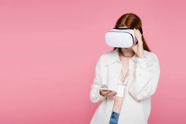 Mujer pelirroja usando auriculares de realidad virtual y usando un teléfono inteligente aislado en rosa - foto de stock